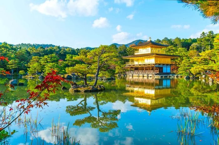 The stunning architecture of Kinkaku-ji (The Golden Pavilion) mirrored in its tranquil lake, surrounded by lush forests and clear blue skies.