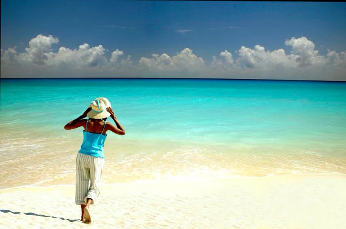 A woman clutches her hat as she strolls towards the ocean on a pristine, white sand beach in Cancún.