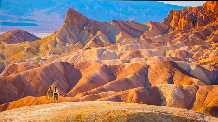 Two individuals stand amid the rugged, undulating terrain of Death Valley National Park, dwarfed by the towering peaks.