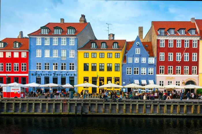 Colorful pastel houses line a canal in Copenhagen, with people enjoying café seating under umbrellas by the waterfront