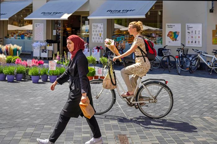 A woman strolls through a square while a cyclist passes by, holding a bouquet of flowers