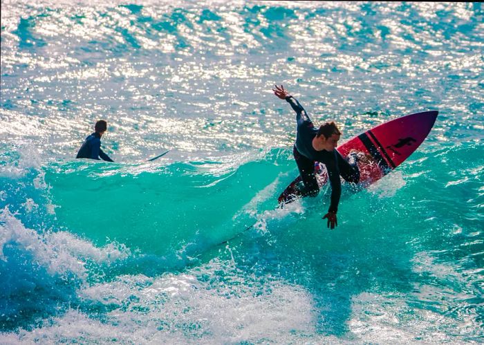 A surfer in a black wetsuit rides a vibrant blue, frothy wave at nearly a 90-degree angle on Praia do Beliche in Portugal, while another surfer in the background scans the sea for the next wave to catch.