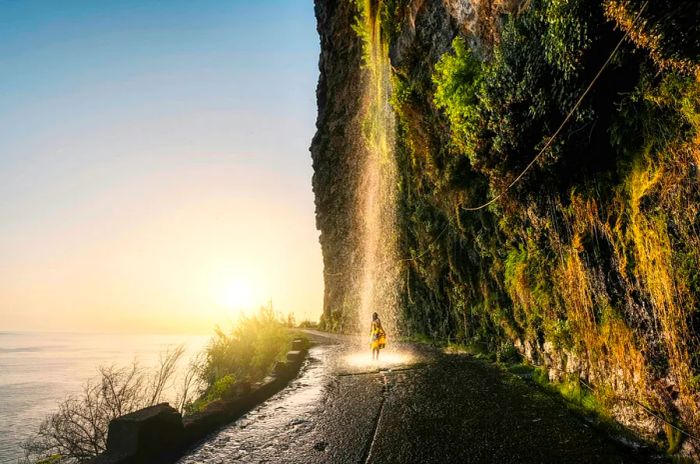 A woman stands beneath a waterfall cascading off a cliff along a road in Madeira, with the sea visible to her left.