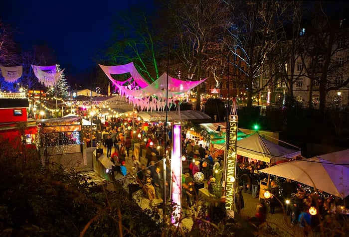 Crowds bustle through the brightly lit Christmas market, flanked by tall, bare trees.