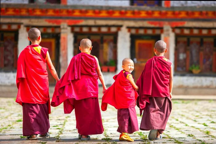 A group of four novice monks in red robes strolls through a courtyard, with one flashing a playful smile at the camera