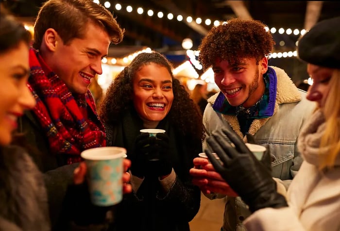 A group of five friends in warm attire smile at each other while holding cups of their drinks.