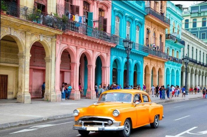 A vintage bright-yellow American car cruising past the vividly painted buildings in Havana Vieja, Cuba.