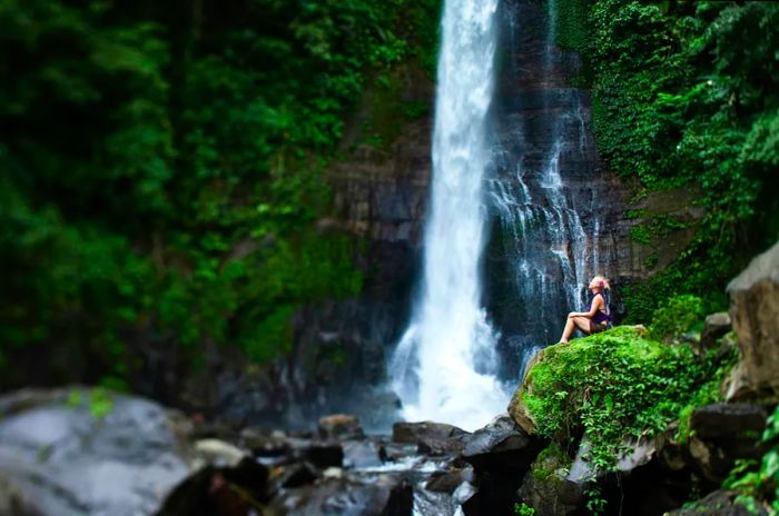 A woman relaxes near a waterfall in Bali, surrounded by lush green forest