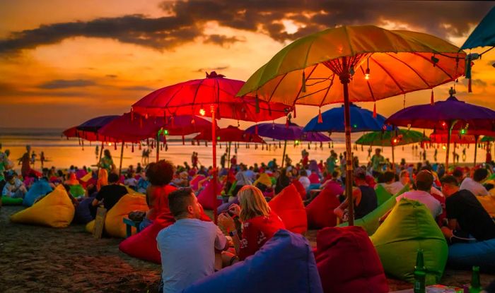 Tourists enjoy a relaxing evening on colorful bean bags under umbrellas while watching the sunset at Denpasar beach, Bali.