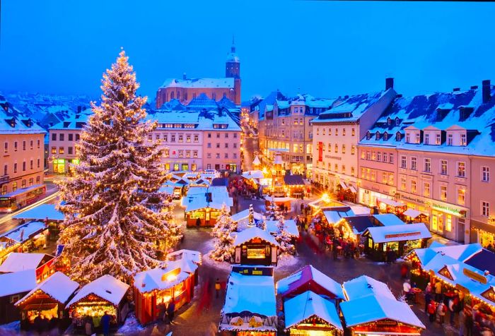 A vibrant Christmas market bustling with activity, featuring a grand Christmas tree at its center, surrounded by the city's architecture illuminated at night.