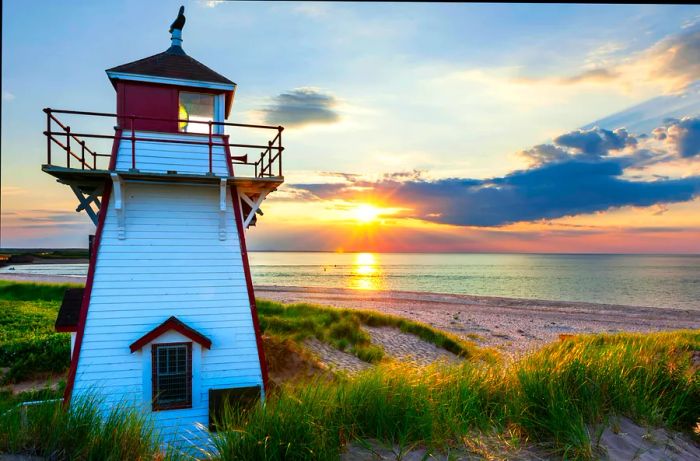 A bright white wooden lighthouse stands prominently in the foreground as the sun sets over a tranquil sea beyond.