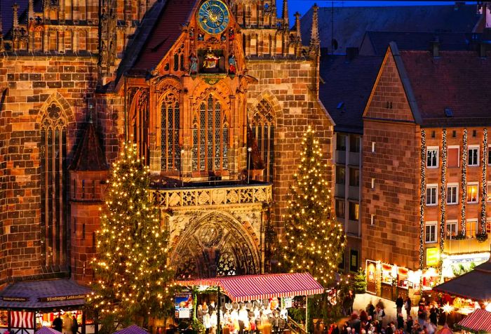 A beautifully adorned Gothic cathedral stands tall, flanked by impressive Christmas trees at its entrance, with festive shops bustling in the main square.