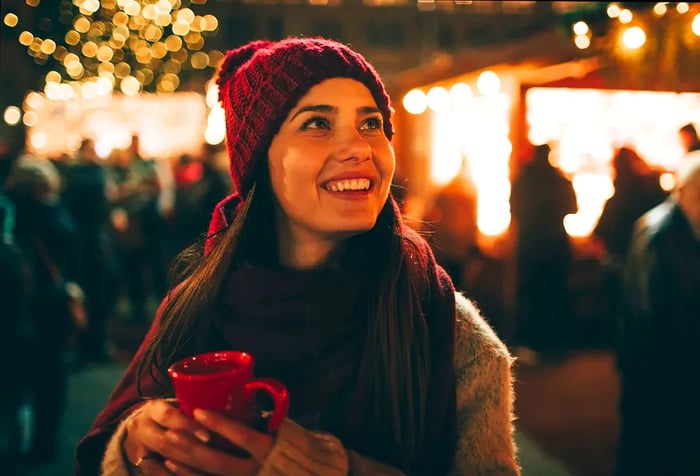 A woman bundled up in winter attire, joyfully holding a red mug and gazing upwards with a smile.