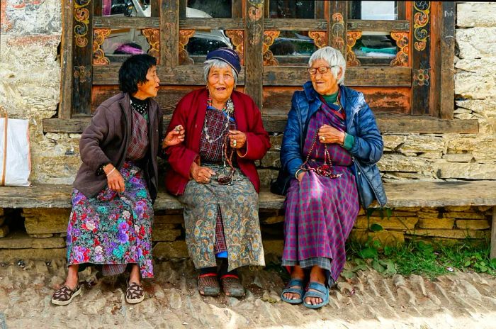 Three women gather in front of a traditional house, chatting and laughing, in Mongar, Bhutan