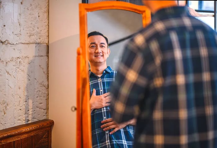 A man adjusts his flannel shirt while looking at his reflection in the mirror.