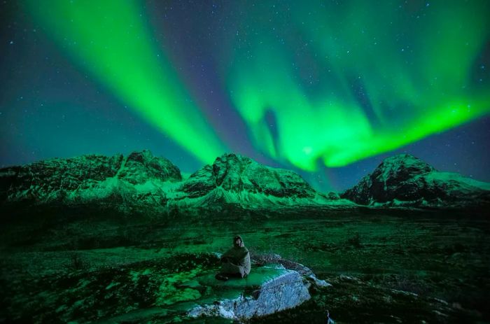 A person sits on a rock in the remote wilderness, gazing up at the enchanting green glow of the Northern Lights.