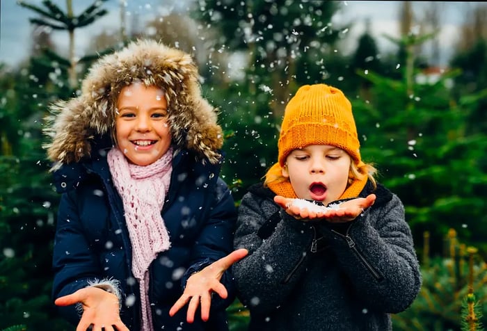 A cheerful child in a parka extends their hands to catch snowflakes blown from the hands of another child in a yellow knitted cap.