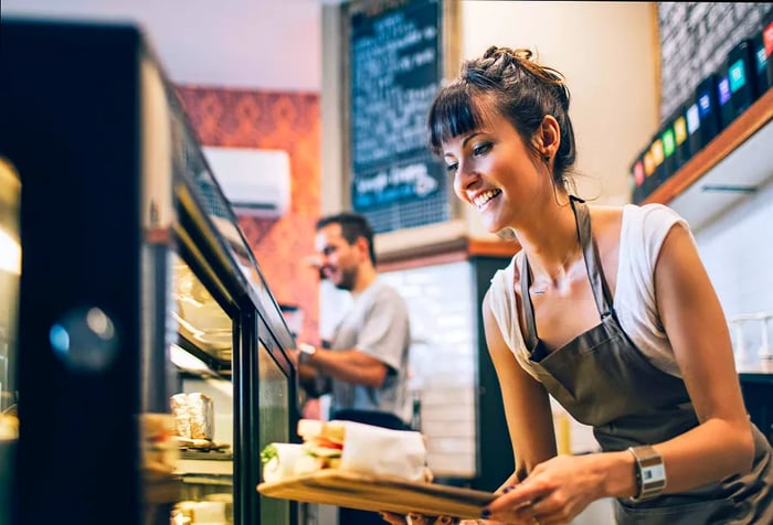 A brunette woman in an apron arranges sandwiches on the display counter of a restaurant.