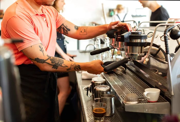 A skilled barista showcases their tattooed arm while expertly crafting a flavorful cup of coffee at the espresso machine.