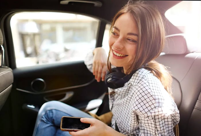 A brunette woman in a white checkered shirt beams as she relaxes in the backseat of a car.