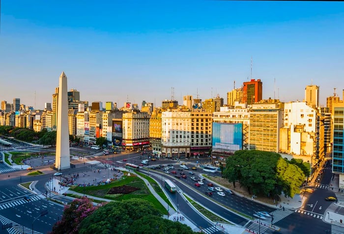 A white stone tower monument stands at the crossroads of busy avenues flanked by tall, compact buildings.