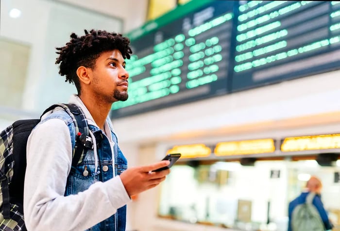 A young man in a denim vest and backpack stands at the airport, using his smartphone to check the flight schedules on the departure board.