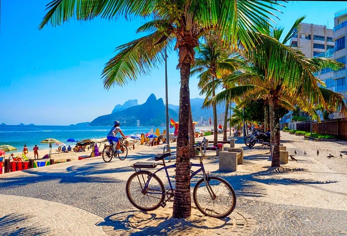 A seaside promenade lined with benches and coconut trees, overlooking a bustling beach.