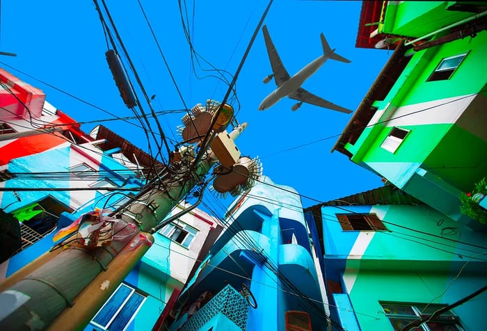 An airplane soaring through the blue sky above a backdrop of vibrant buildings.