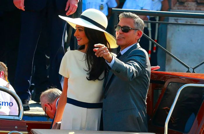 George and Amal Clooney arriving at their civil ceremony in Venice by boat; she dons a cream trouser suit and wide-brimmed hat while he sports a dark grey suit.