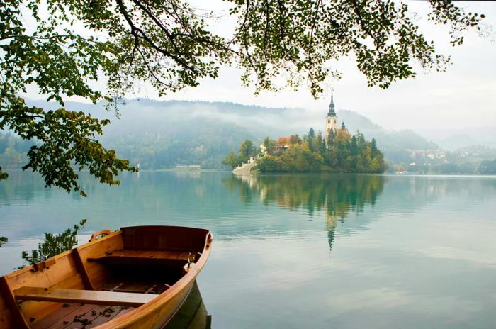 Vibrant rowing boats line the shores of Lake Bled, framed by autumn-colored trees, with Bled Island and the Church of Mary the Queen gracefully positioned in the background.