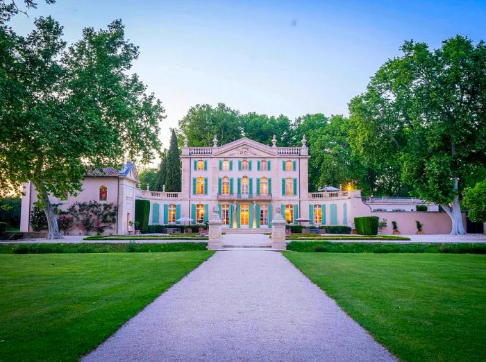 A gravel path winds up to a magnificent stone French chateau featuring bright turquoise shutters on its windows, surrounded by lush greenery.