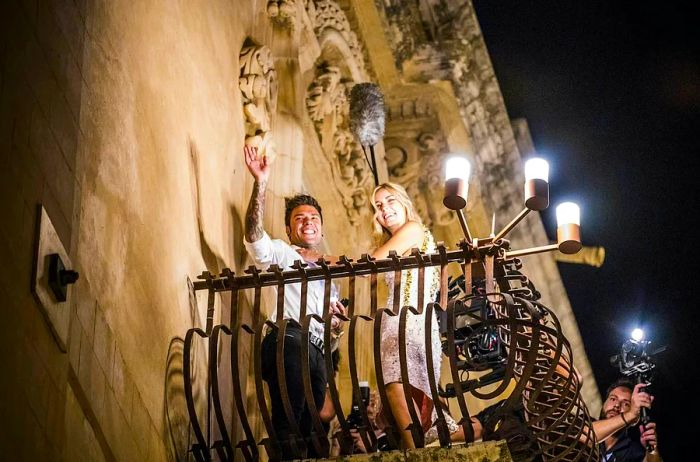 Gazing up at Chiara Ferragni and Fedez as they stand on the ornate wrought-iron balcony of a lavish stone palazzo, smiling and waving down at the camera.