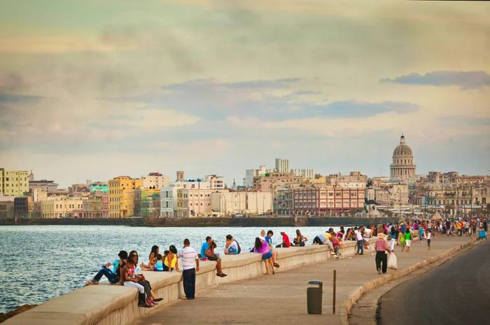 As evening falls, people gather along the seawall to watch the sunset, with a stunning skyline featuring a prominent domed building behind them.