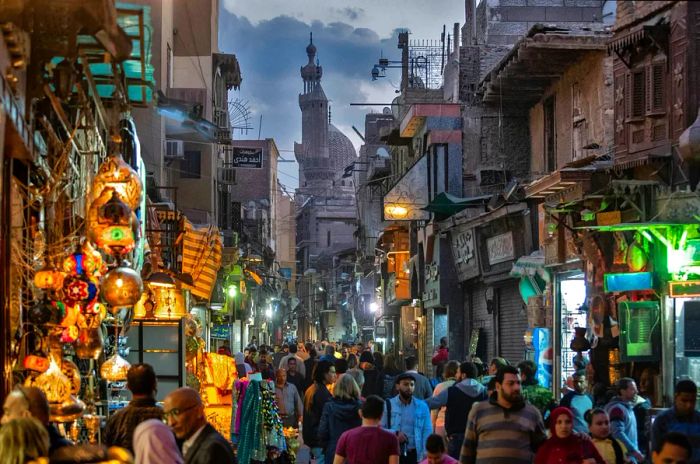 A bustling pedestrian street in the late afternoon, with a large mosque visible at one end.