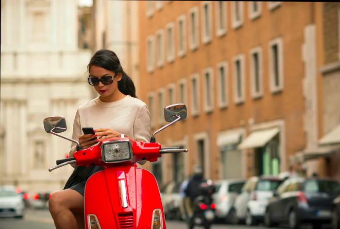 A young woman checks her phone while sitting on a parked red moped in Piazza del Popolo, Rome, Italy.