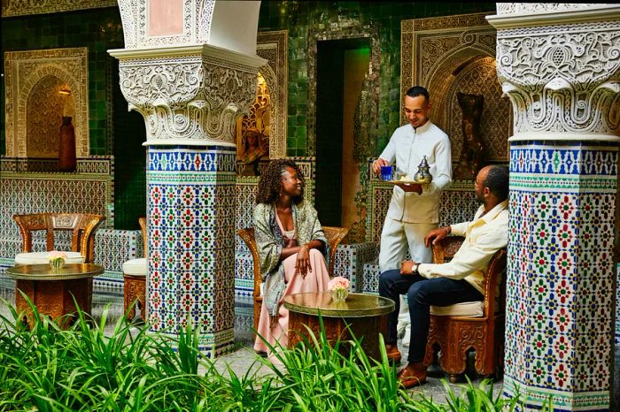 A wide shot of a cheerful couple being served tea by a waiter in the luxurious courtyard of a hotel during their vacation.