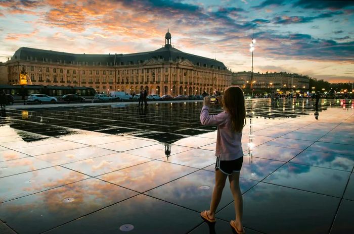 A young girl captures the beauty of the iconic Bordeaux water mirror, with Place de la Bourse illuminated in the background at sunset.