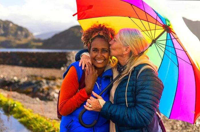 A close-up of a same-sex female couple sharing a laugh under a vibrant umbrella. The woman on the right kisses her partner on the side of the head as they stand by Loch Torridon near Diabaig in the stunning Northwest Highlands of Scotland.