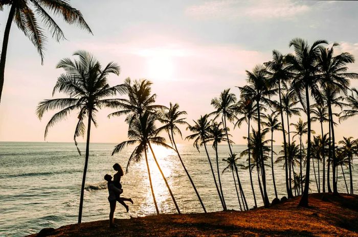 A loving couple enjoys a sunset over the sea, surrounded by palm trees. A boy and girl experience the magic of a sunset. Newlyweds bask in the beauty of the islands.