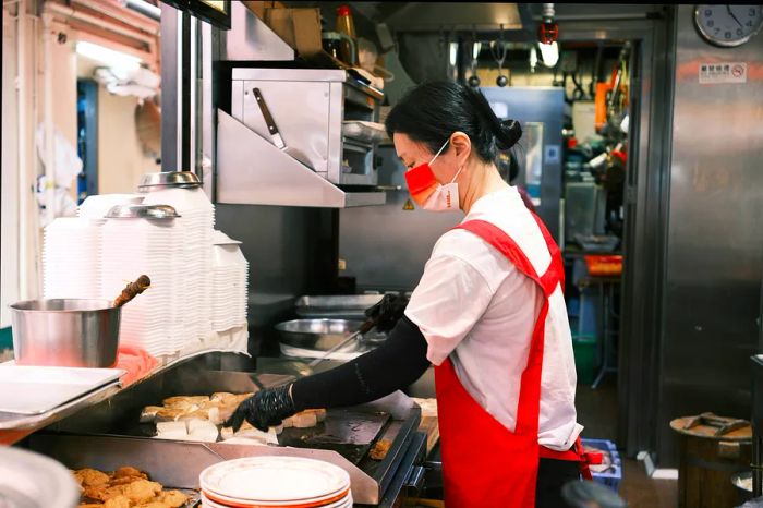 A woman skillfully prepares fried tofu on a griddle in a bustling industrial kitchen.