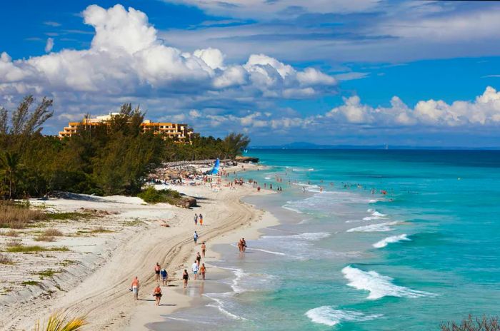 A stunning white-sand beach with a hotel in the background, where many people congregate on the sand near the building.