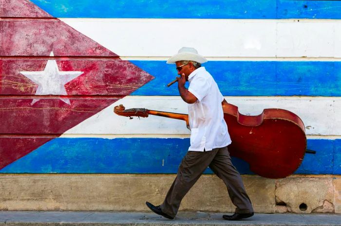 A man with a large bass instrument smokes a cigar as he strolls past a Cuban flag mural on a wall