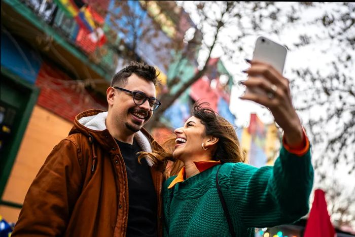 A joyful couple snaps a selfie beneath a tree in Buenos Aires, Argentina, with vibrant painted houses in the backdrop.