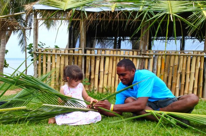 A Fijian man teaches a young girl how to weave a basket using palm fronds.