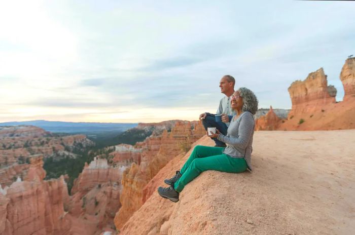 An older couple pauses during their hike to take in the breathtaking view from a Utah overlook, sitting on the cliff's edge and soaking in the scenic beauty of the mountains and canyon before them.