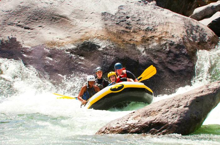 A group of four helmeted adventurers navigate a white-water raft down the Río Cangrejal in Honduras, surrounded by massive boulders.