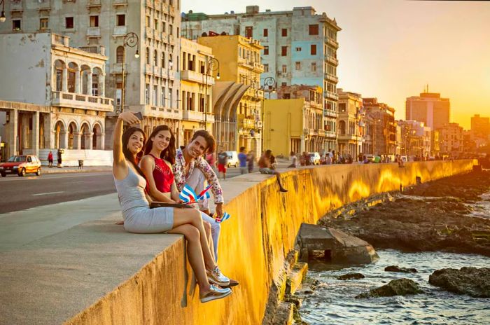 A group of friends relax on a coastal wall, dangling their legs over the water as they take a selfie.