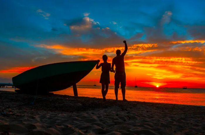 A couple silhouetted by the stunning red and orange hues of a sunset beside a boat in Roatán, Honduras, Central America.