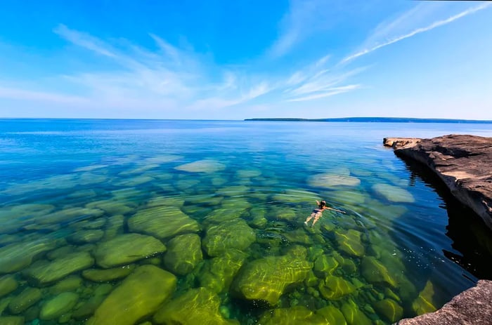 A girl enjoys a swim in Lake Superior in Michigan's upper peninsula, where rocks are visible through the crystal-clear waters. Pictured Rocks National Lakeshore can be seen in the distance.