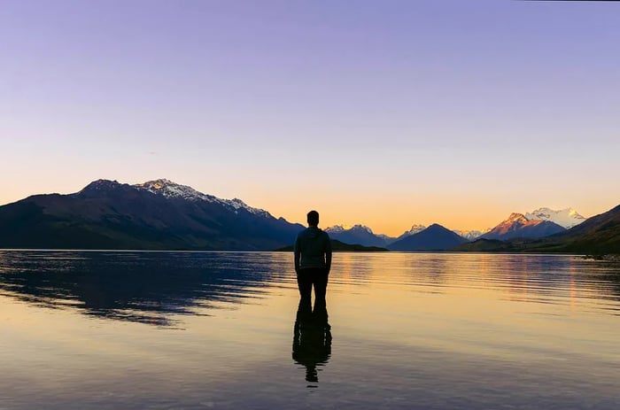 A man gazes at the breathtaking sunset behind the snow-capped peaks while standing in Lake Wakatipu, New Zealand.